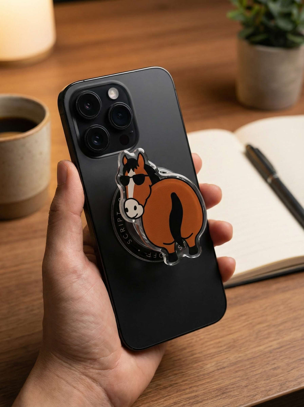 Hand holding black iPhone with OFF SCRIPT Flattery Little Horse MagSafe magnetic phone grip attached, on a wooden desk background